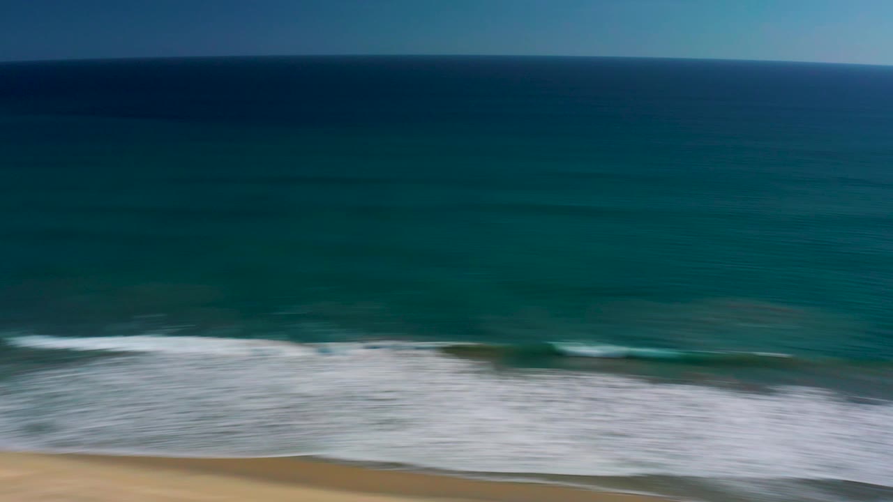 Aerial frame over the San Jose del Cabo coast with dark blue ocean at left, white surf reaching the sandy beach, bright water glare, and no visible people.