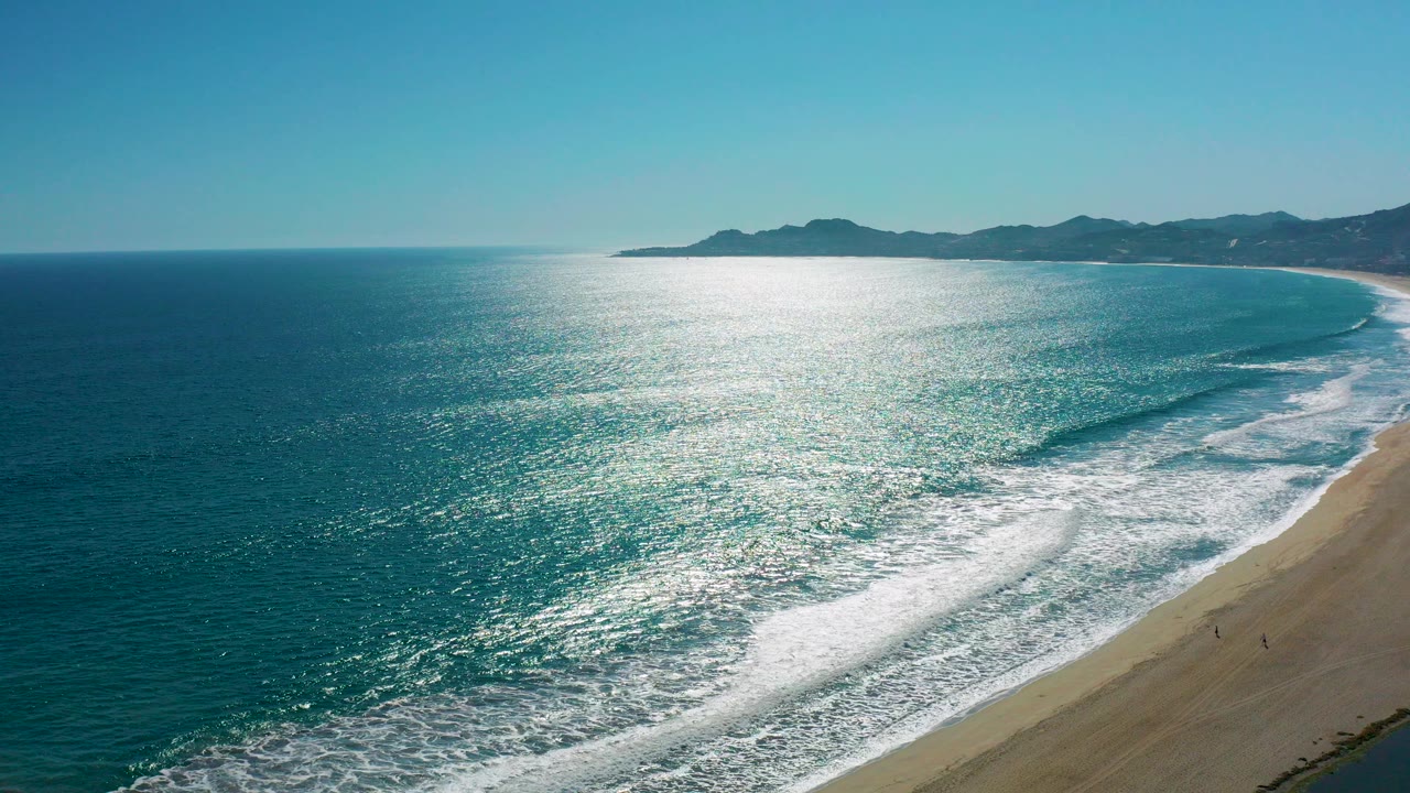 A high aerial view over San Jose del Cabo coastline shows sunlit blue-green water, white surf foam, sandy beach on the right, and low hills on the horizon.
