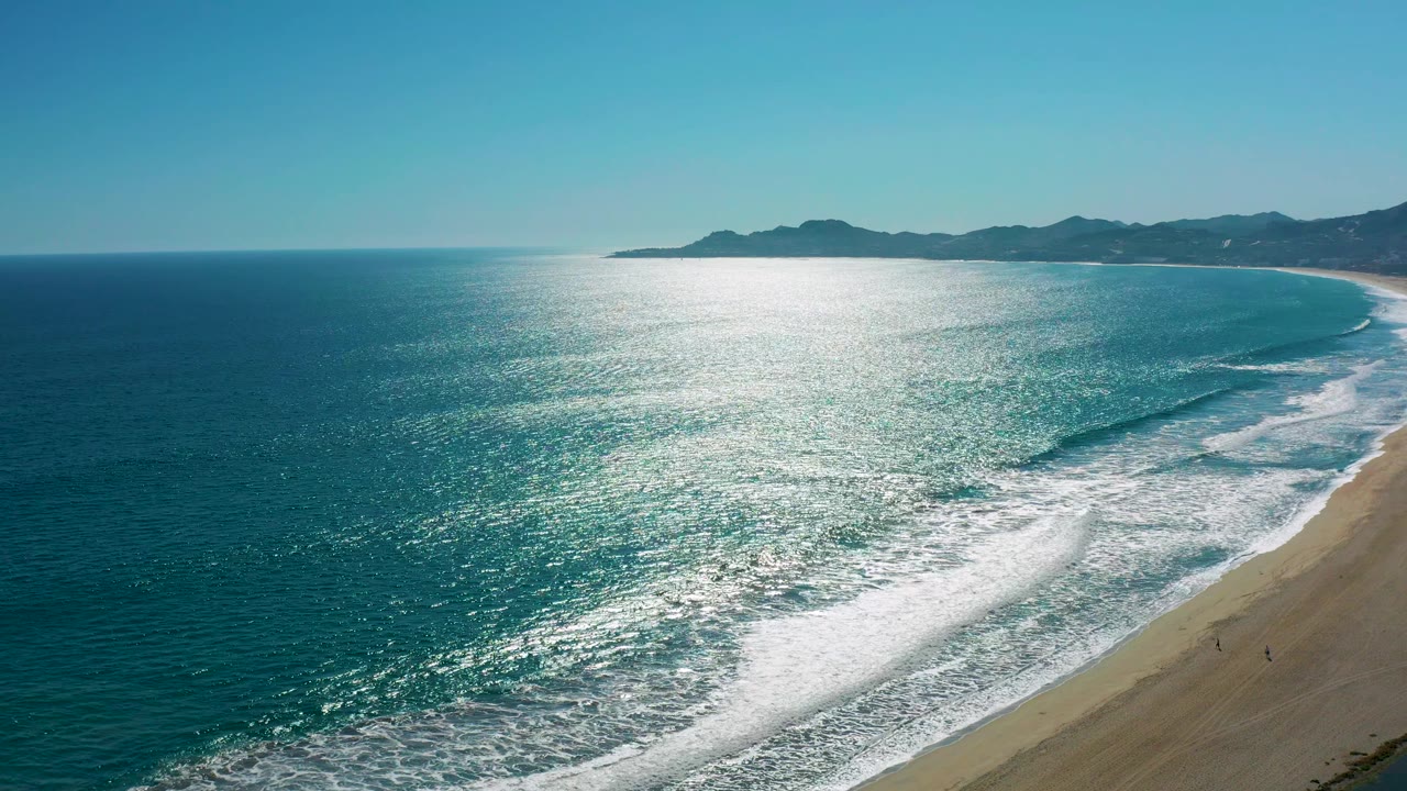 A wide coastal frame shows bright glare across the water, rolling surf near the beach, and dark distant hills beyond the San Jose del Cabo shoreline.