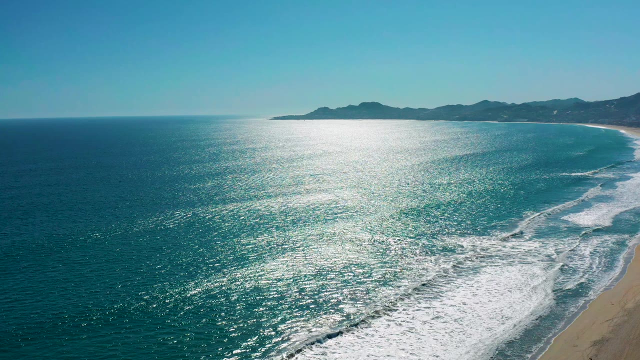 San Jose del Cabo shoreline seen from above, with glittering blue water filling the frame, a narrow strip of sandy beach on the right, and no visible people.