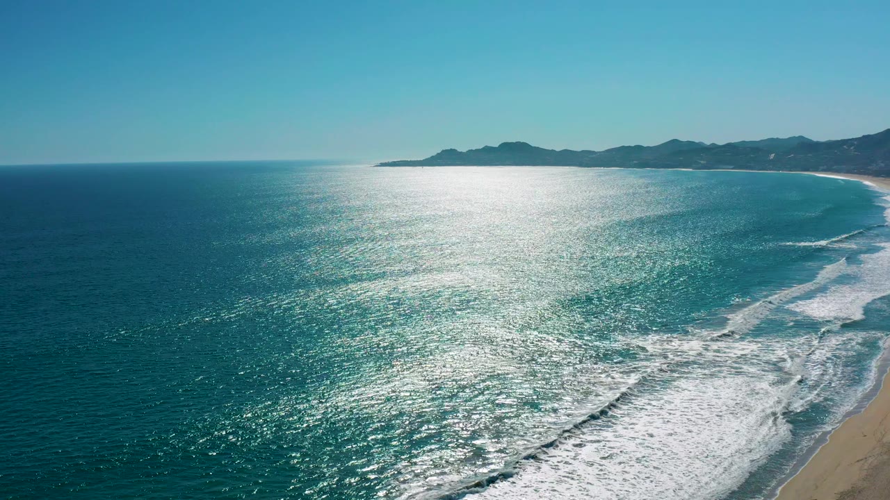 High coastal view in San Jose del Cabo with white foam bands advancing across turquoise water, sand along the lower right, distant hills, and no visible people.