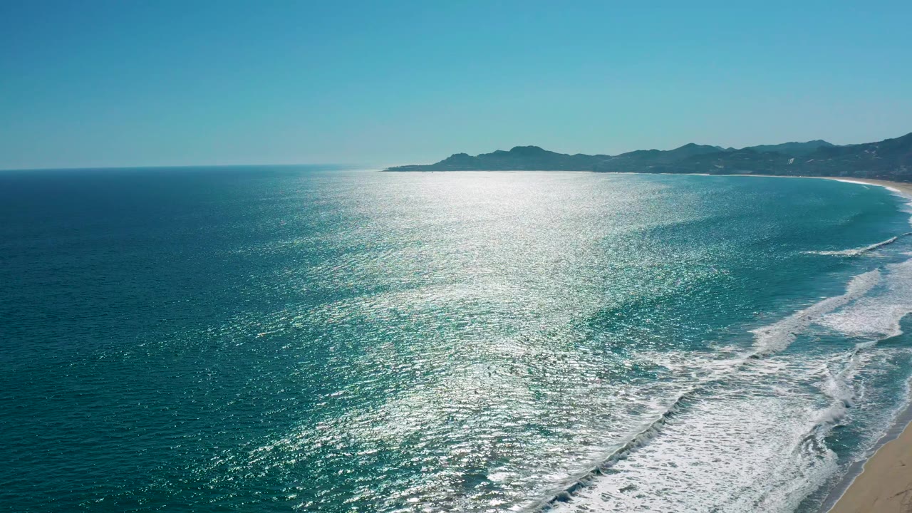 Wide overhead view of the San Jose del Cabo coast with a broad patch of sunlit ocean, white surf curling near the right edge, sandy beach, and no visible people.