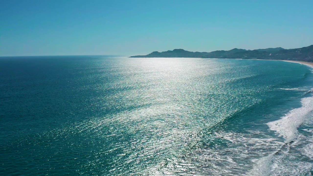 Aerial frame of San Jose del Cabo coastline where a band of white foam curls beside the sandy shore under a clear blue sky, with no visible people.