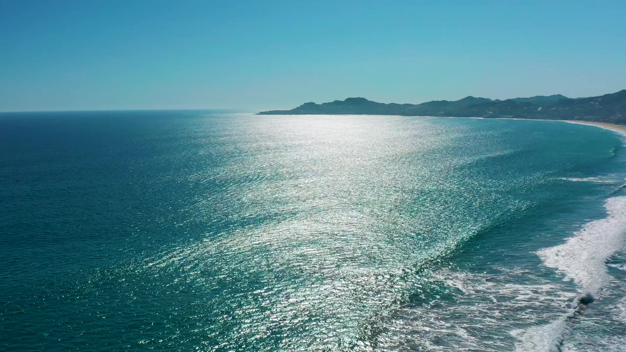 High aerial view of San Jose del Cabo ocean with the silver reflection centered on the water, the surf pushed farther right, low mountains in back, and no visible people.