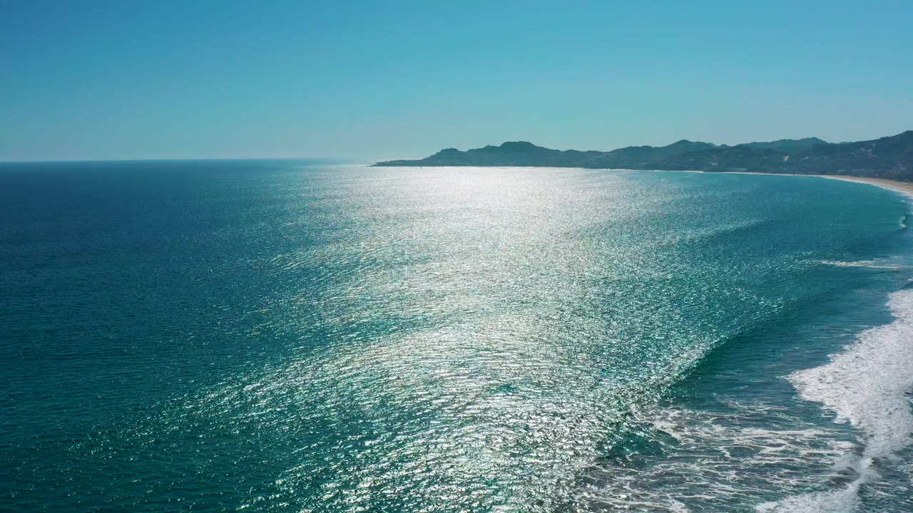 Aerial view over San Jose del Cabo water where the shoreline curves along the right side, white foam gathers near the beach, and no visible people appear in the scene.