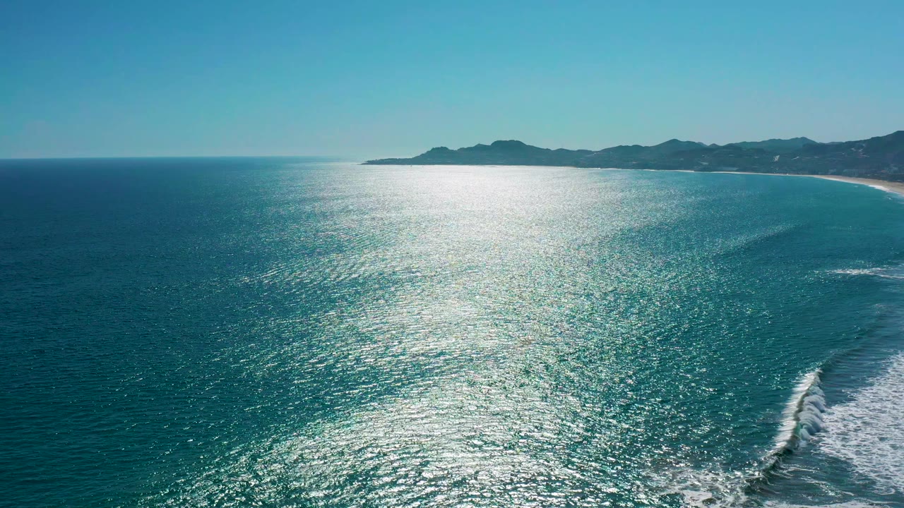 Wide aerial frame of San Jose del Cabo coastline, with sparkling blue water across the foreground, a white wave line nearer the lower right, mountains on the horizon, and no visible people.