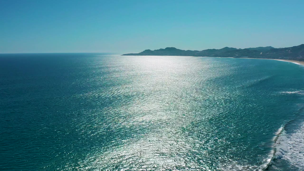 Aerial view of San Jose del Cabo with the sun glare centered over textured blue water, a pale sandy shoreline to the right, distant mountains, and no visible people.