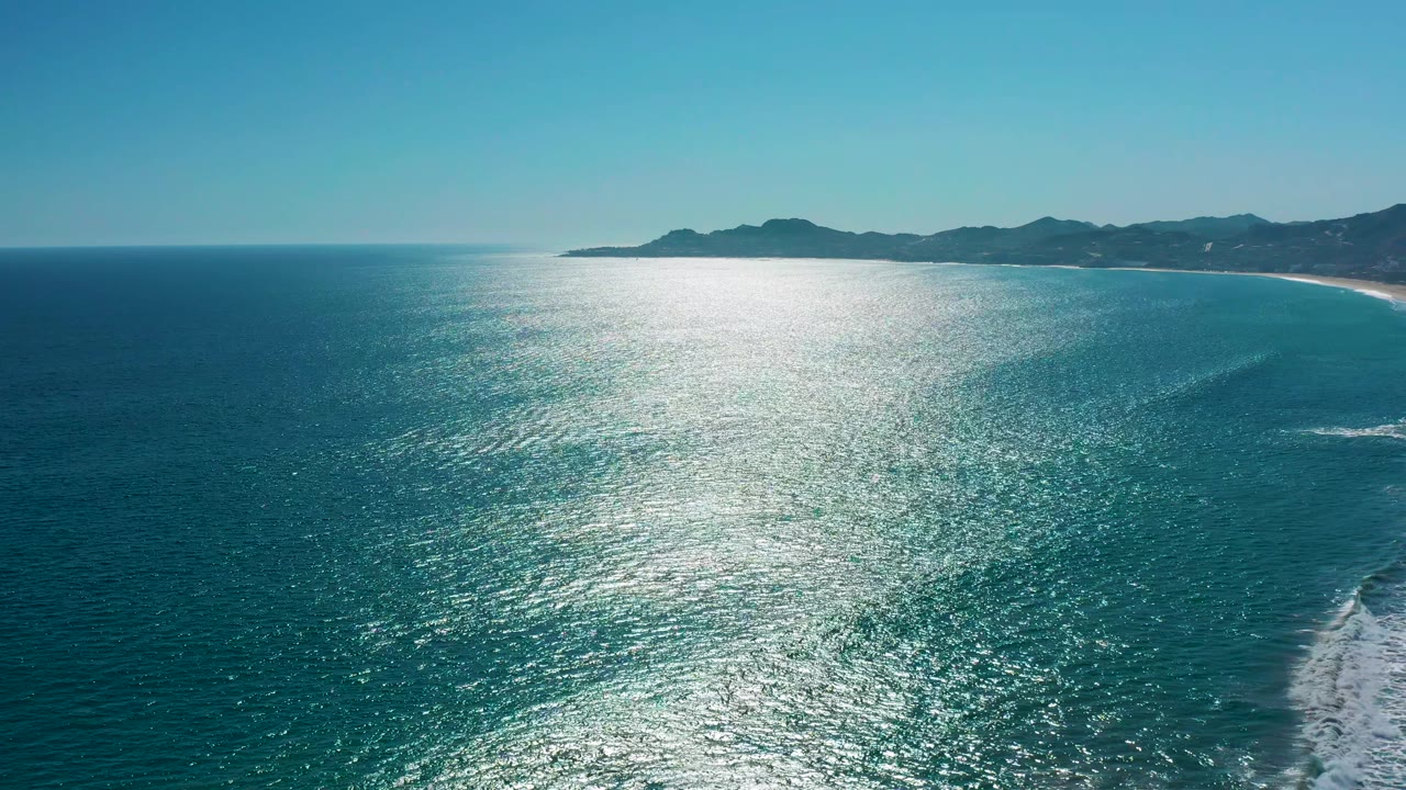 Aerial coastal view over San Jose del Cabo water, with strong sunlight glittering across the sea, a thin sandy beach along the right, mountain silhouettes in back, and no visible people.