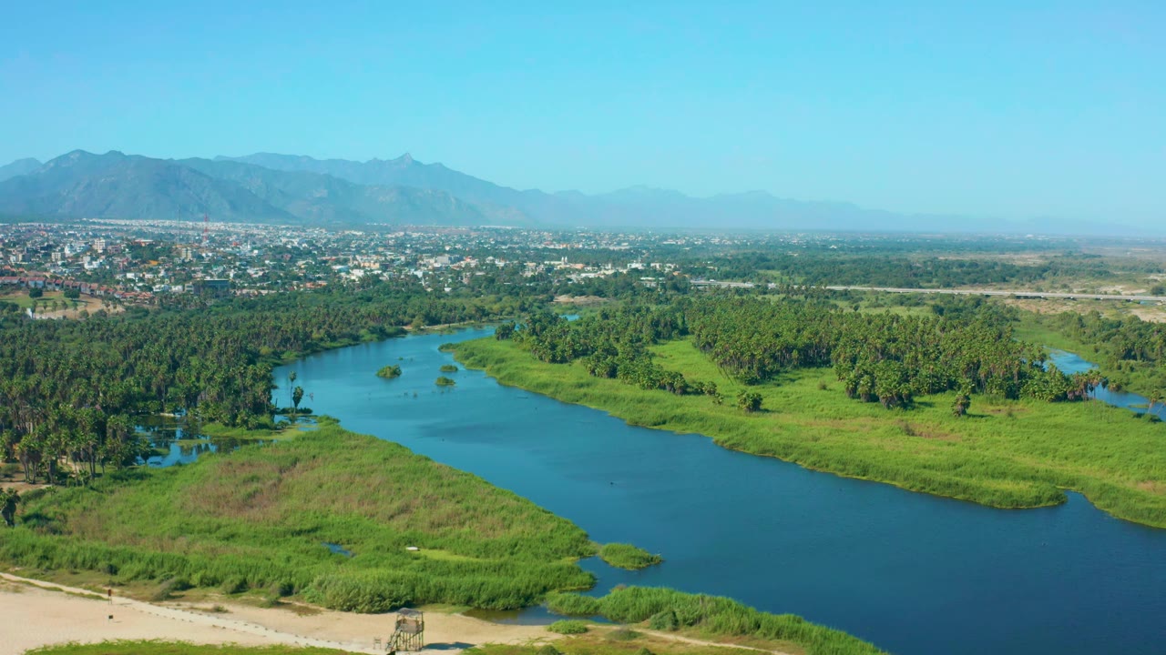 Aerial frame over San Jose Del Cabo, Mexico, showing the river bend slightly farther across the foreground, with thick green tree cover, town buildings, hazy mountains, and no visible people.