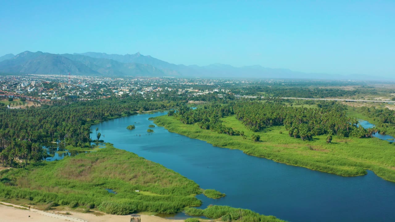 Aerial view in San Jose Del Cabo, Mexico, with the blue river running along dense green trees and open grass, distant town blocks below the mountain ridge, and no visible people.