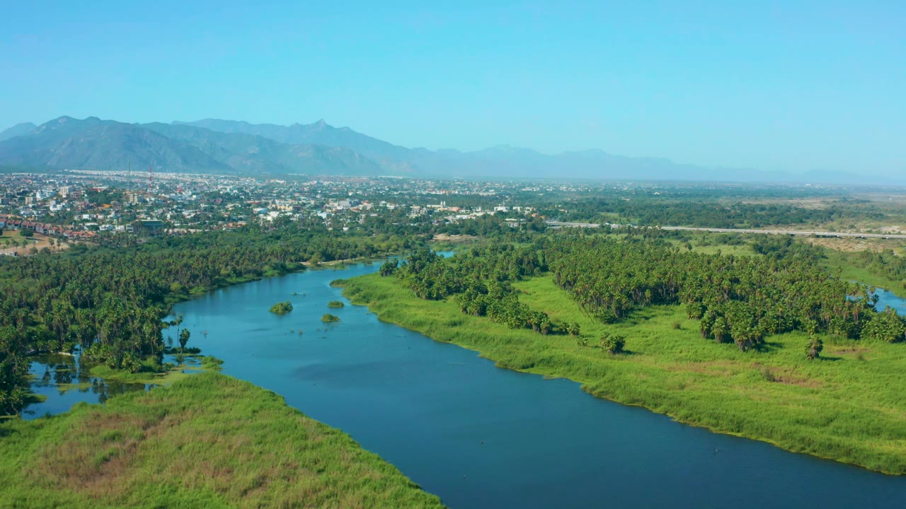At one second, the San Jose del Cabo aerial holds the winding river in the foreground, green banks and trees in the middle distance, and town buildings below hazy mountains, no visible people.