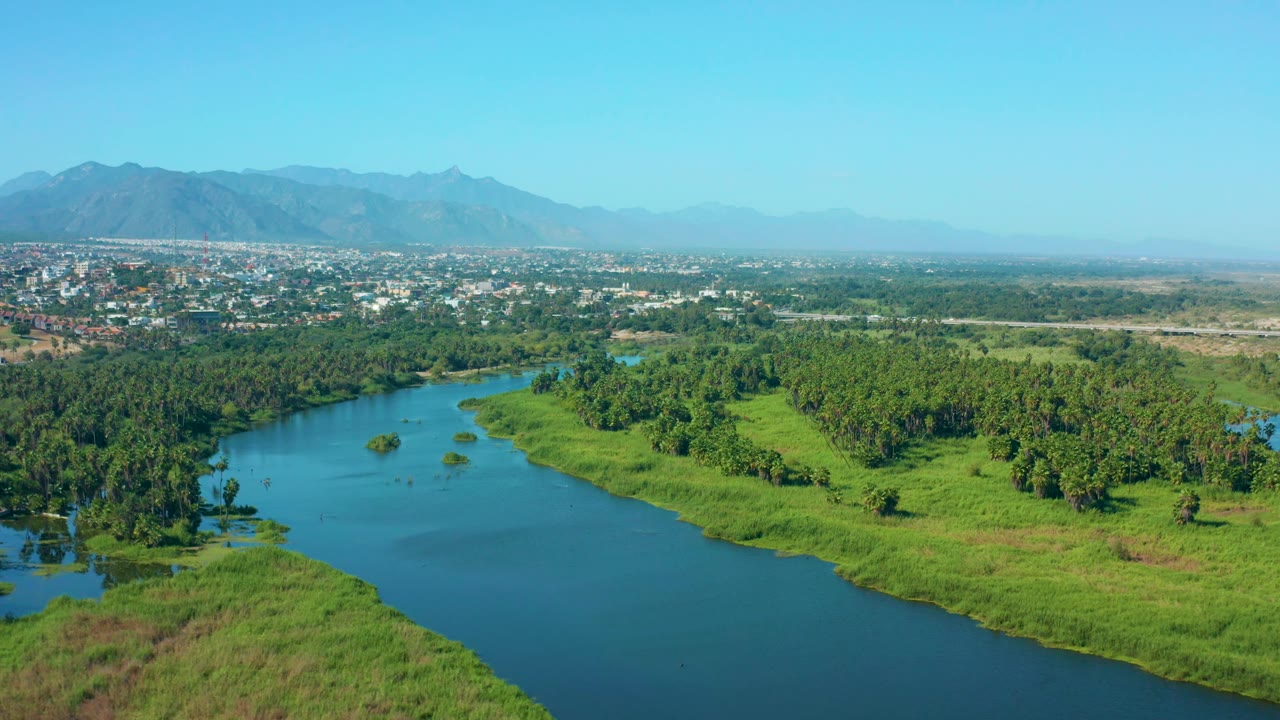 At three seconds, the aerial view shows a broad river bend bordered by green wetland grass and tree lines, with San Jose del Cabo and mountains across the horizon, no visible people.