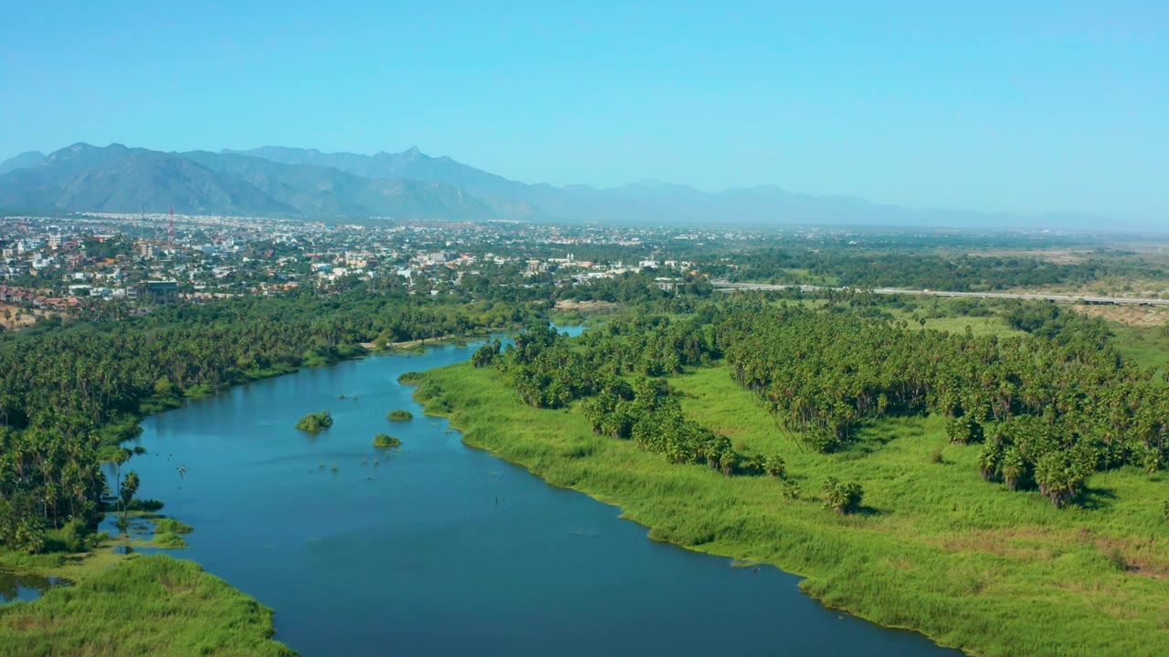 Aerial river bend bordered by grass and trees with town and mountains beyond.
