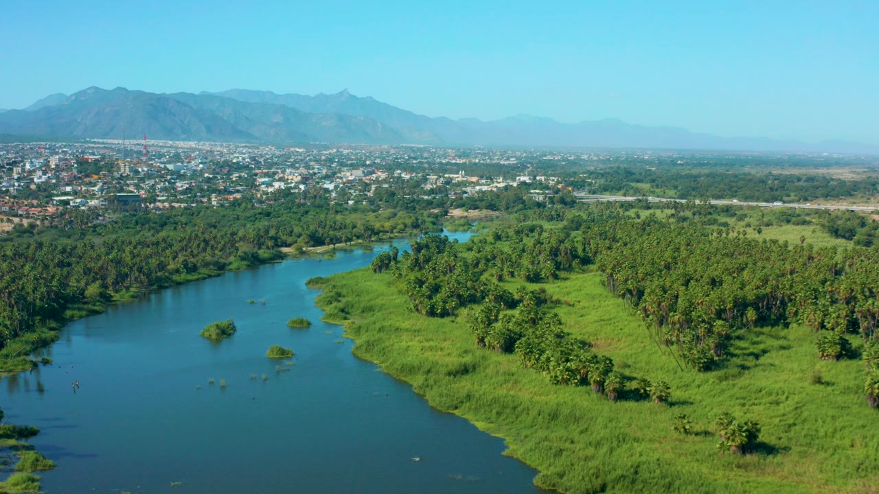 Opening aerial frame over San Jose Del Cabo, Mexico, showing a blue river beside dense green vegetation, distant city buildings, hazy mountains, and no visible people.