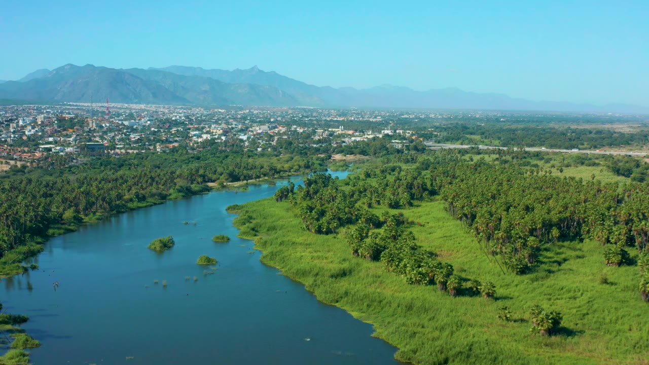 Aerial view in San Jose Del Cabo, Mexico, with the river bending through bright green banks, tree cover filling the right side, city buildings beyond, mountains on the horizon, and no visible people.
