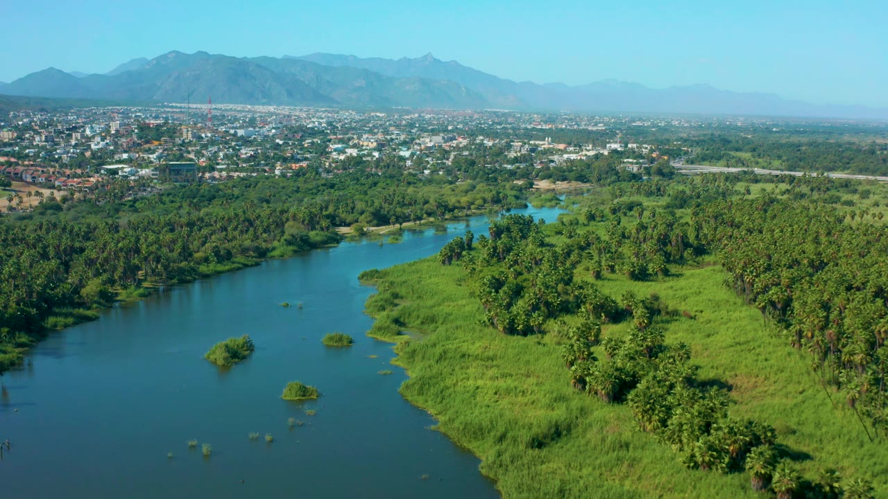 Aerial view over San Jose Del Cabo, Mexico with the river curving through green banks, small islets in the water, dense trees, a town strip, hazy mountains, and no visible people.