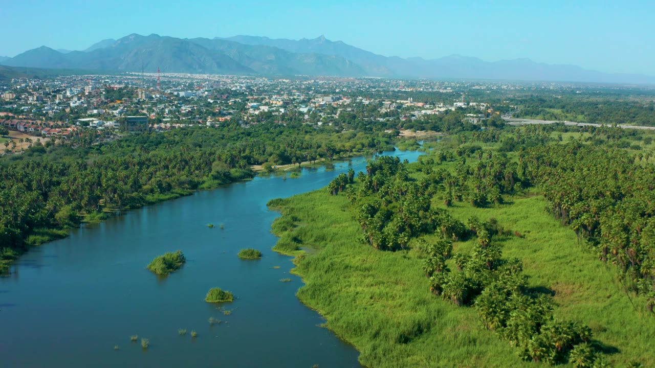 High aerial frame from San Jose Del Cabo, Mexico with open blue water in the foreground, green riverbank plants and trees to the right, town structures in the distance, mountains, and no visible people.