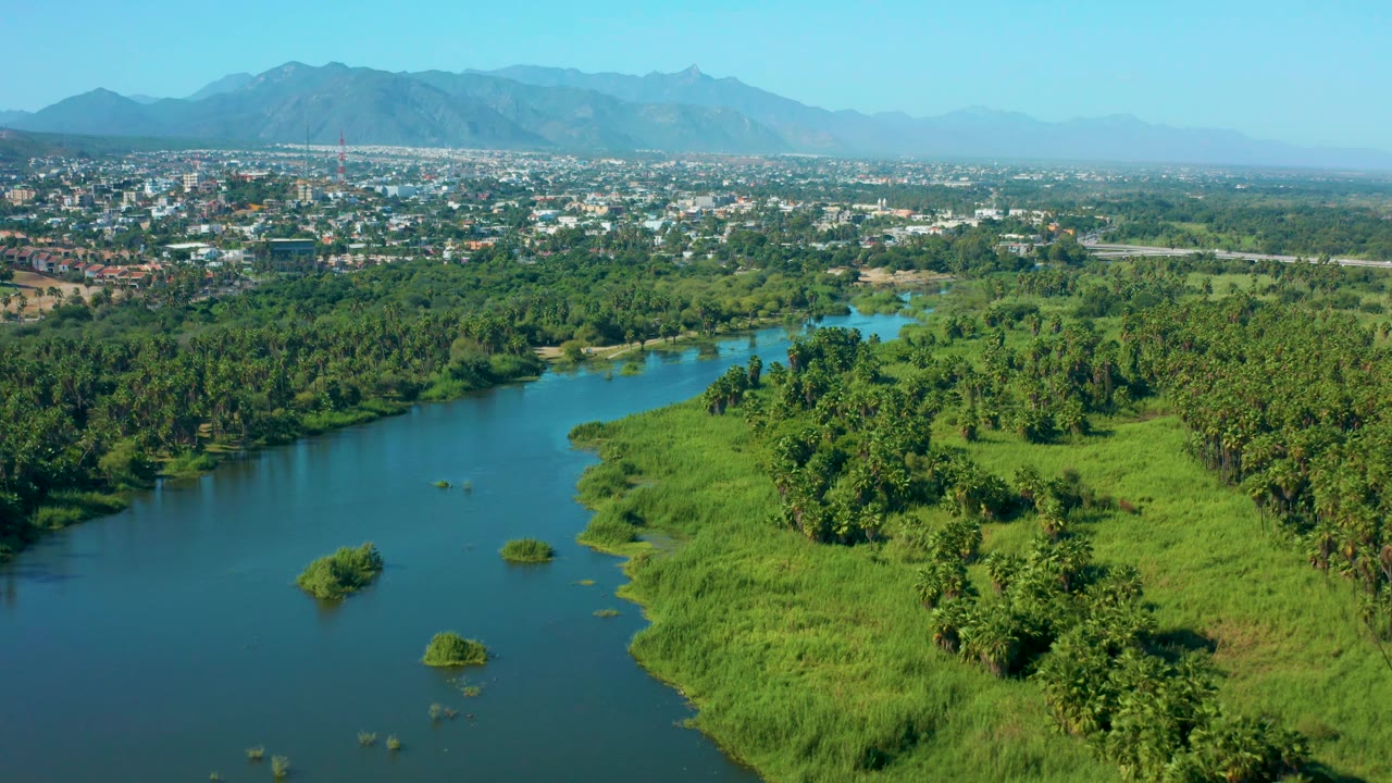 Aerial frame over San Jose Del Cabo, Mexico showing the river channel along the lower left, bright green vegetation across the bank, dense trees in the midground, town development, mountains, and no visible people.