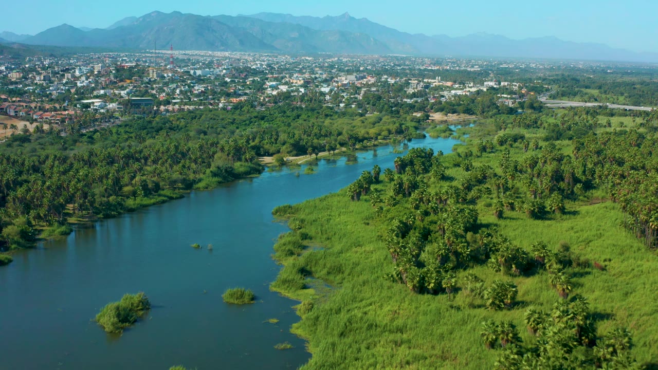 Aerial view over San Jose del Cabo with the river curving through thick green vegetation, small shrubs in the water, town structures in the distance, mountains, and no visible people.