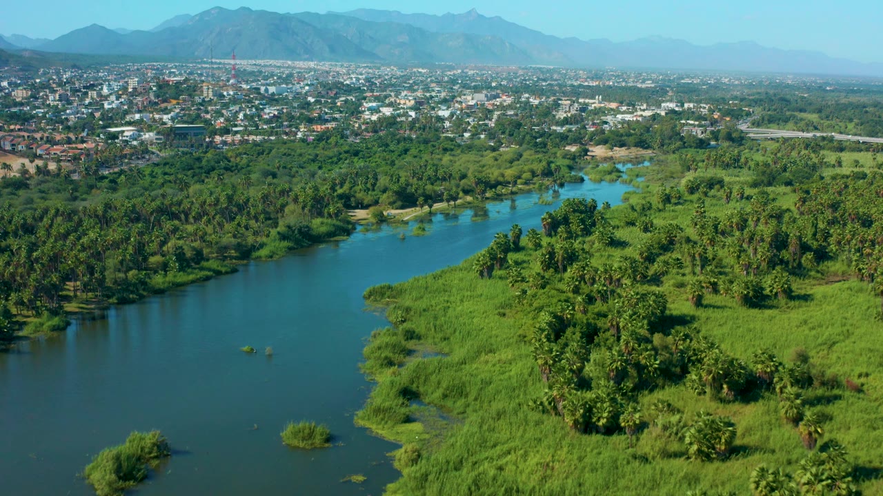 Aerial San Jose del Cabo view showing the river channel set lower in the frame, dense trees and grasslands across the middle, the town beyond, hazy mountains, and no visible people.