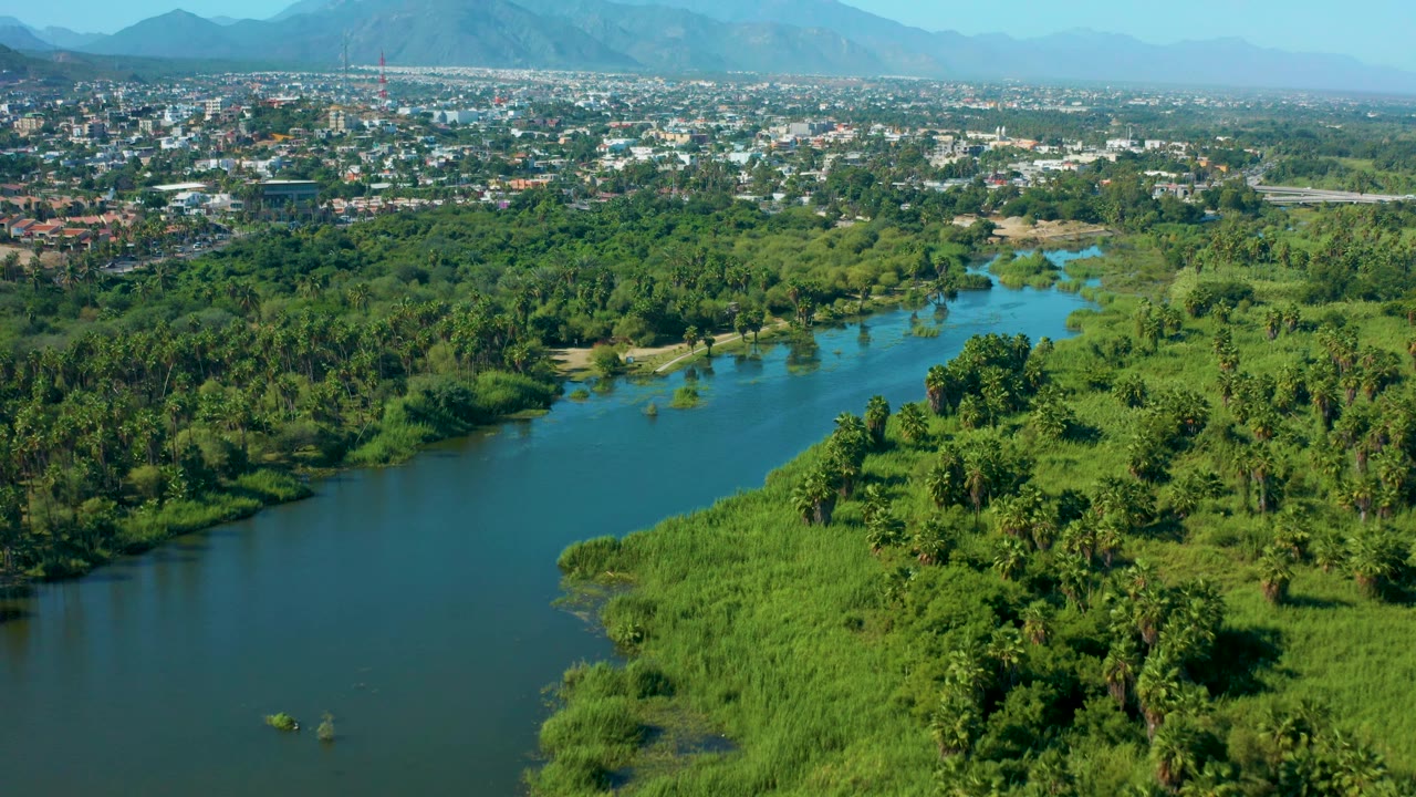 San Jose Del Cabo aerial frame showing a wide river bend, dense palms and grasses in the foreground, a built town strip in the distance, hazy mountain shapes near the horizon, and no visible people.