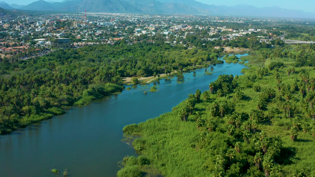 Mid-clip aerial frame over San Jose Del Cabo, Mexico, with open river water on the left, a broad green corridor through the center, town buildings along the top edge, faint mountains, and no visible people.
