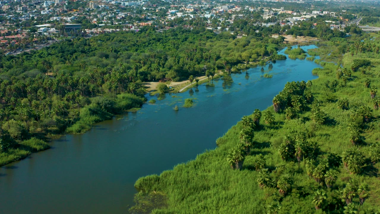 Aerial view in San Jose del Cabo showing a blue-green river bordered by reeds and dense trees, with urban buildings on the far horizon and no visible people.