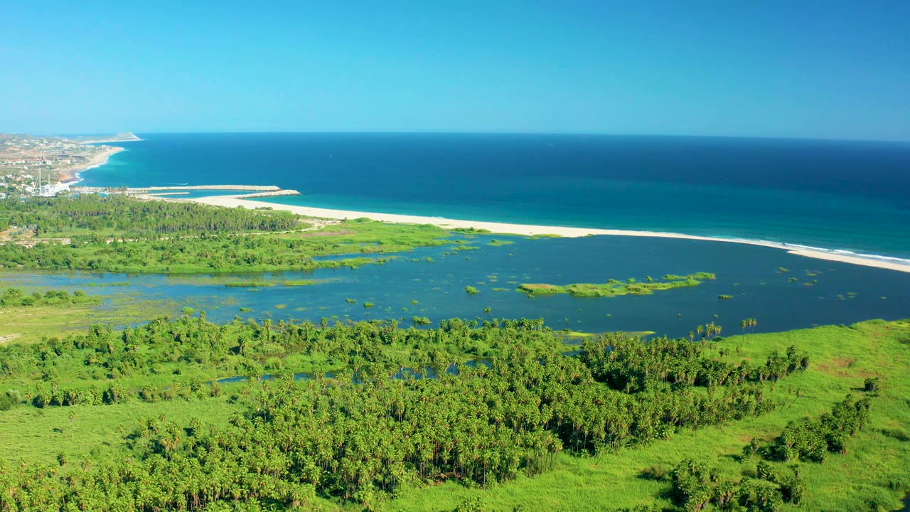 Mid-clip aerial over San Jose del Cabo with green land across the foreground, dark lagoon water through the middle, a bright sand strip, distant surf, blue ocean, and no visible people.