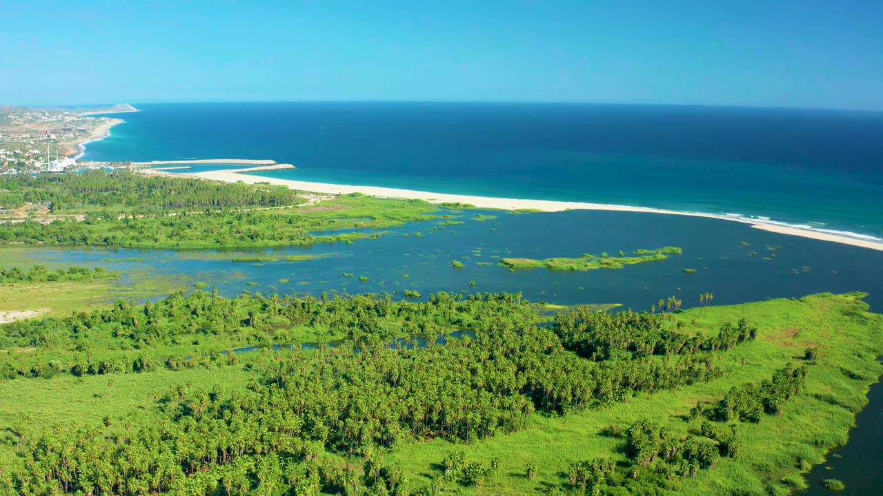 Wide aerial frame in San Jose del Cabo, Mexico, showing foreground wetland vegetation, a dark lagoon, pale sandbar, deep blue ocean, distant coast at left, and no visible people.