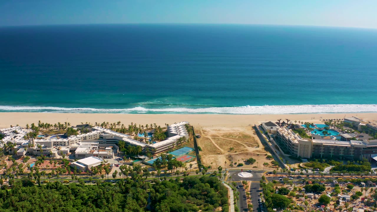 Aerial frame in San Jose del Cabo with the resort complex centered below the beach, white foam advancing toward shore, blue ocean beyond, and no visible people.