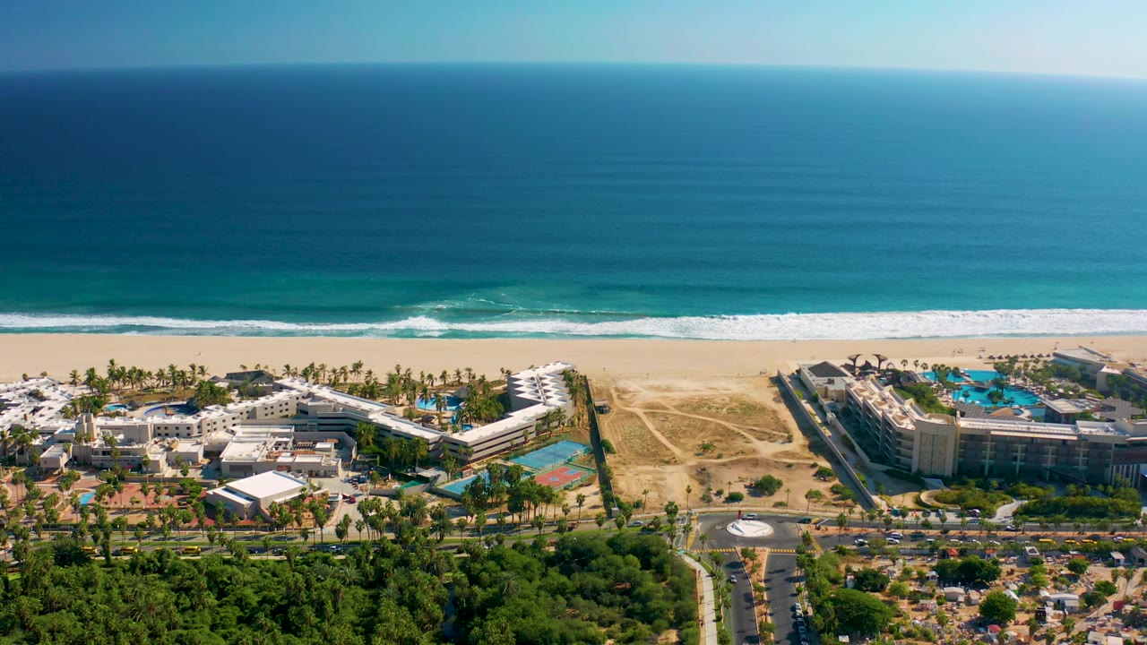 San Jose del Cabo aerial frame showing parallel bands of blue water, white surf, pale sand, resort rooftops, turquoise pool areas, and palms with no visible people.