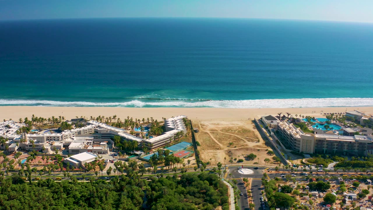 Mid-clip aerial view over San Jose del Cabo with calm blue ocean, a bright white wave band, sandy beach, resort blocks, palms, and roadways with no visible people.