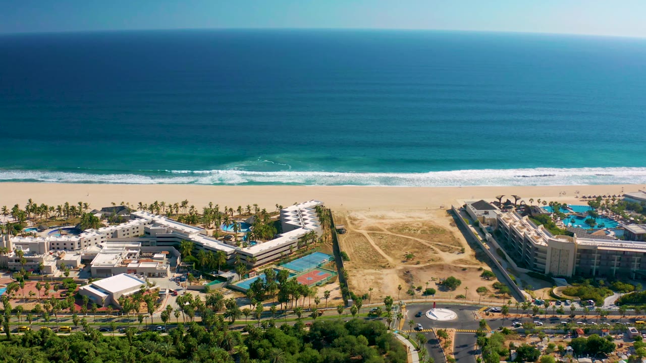Aerial coastline view with ocean horizon, sandy shore, resort roofs, pools, palms, and road.