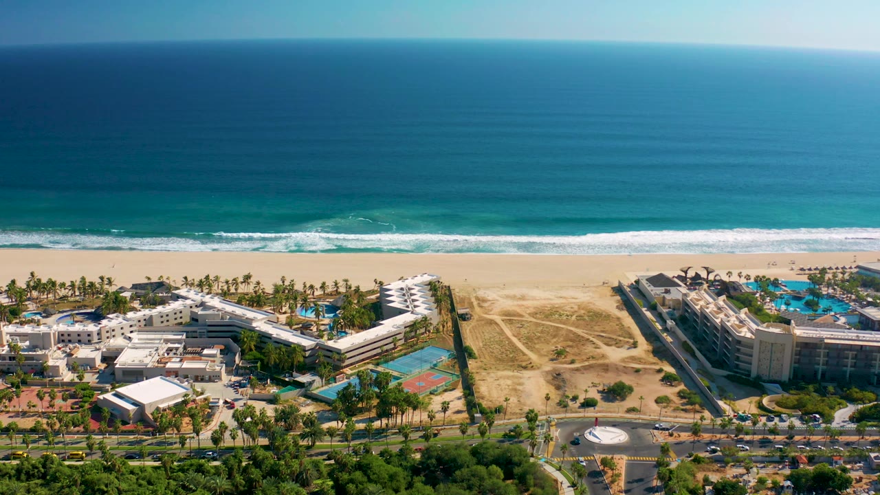 Aerial frame in San Jose del Cabo showing beachfront resort blocks, turquoise pool areas, palm rows, a broad sandy beach, blue ocean bands, and no visible people.