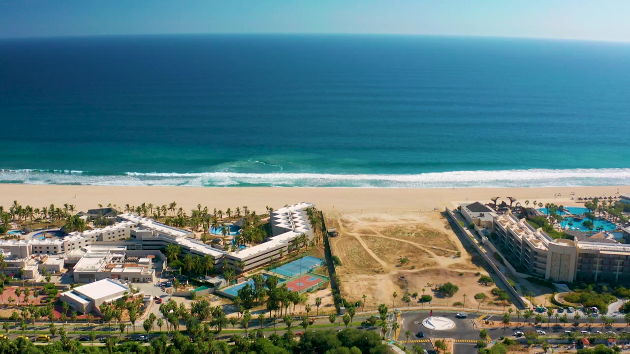 Aerial beach resort view with ocean, surf, pools, palms, road, roundabout, and courts.