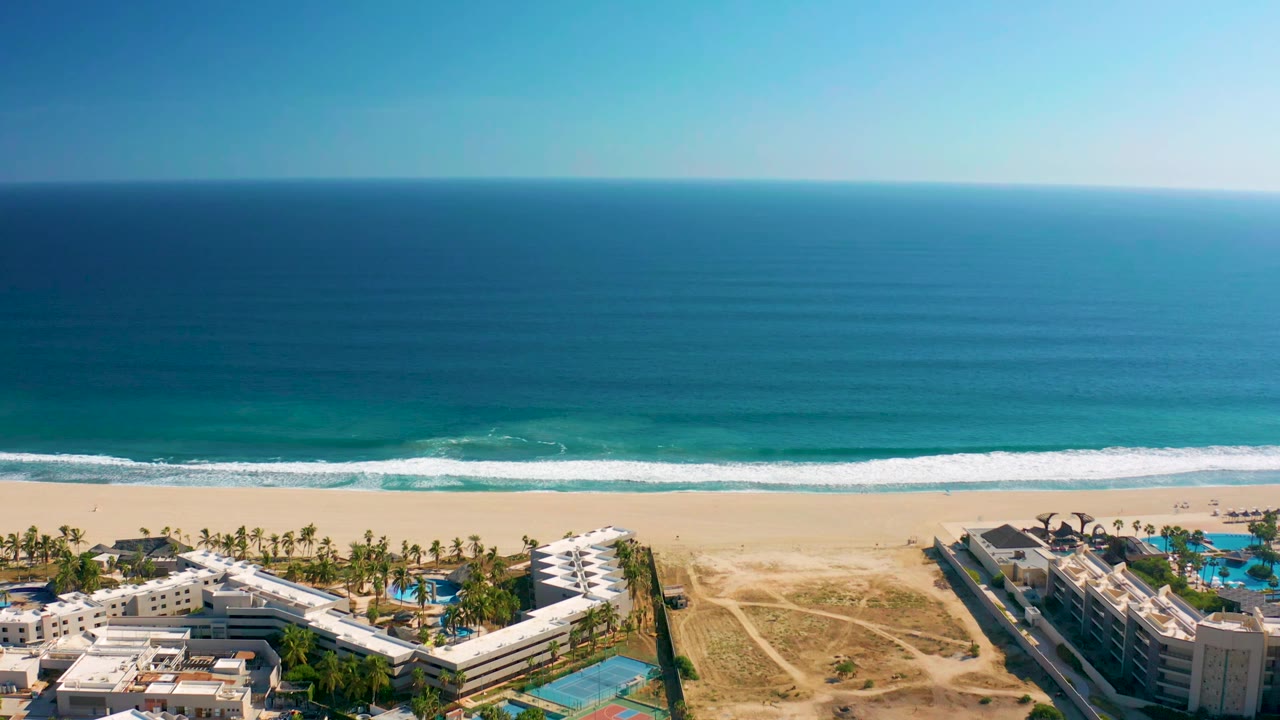 High aerial frame in San Jose del Cabo, Mexico, with beachfront resort rooftops, palm trees, pool areas, pale sand, rolling surf, and no visible people.