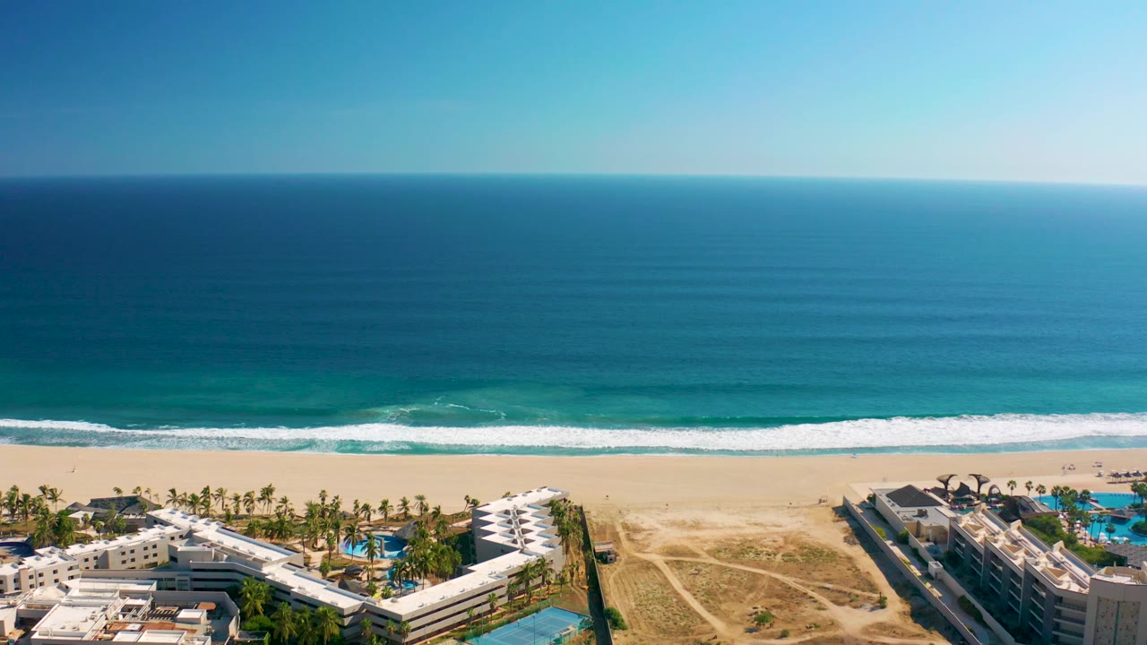 Aerial San Jose del Cabo beach view with a level ocean horizon, bands of turquoise water, white wave foam, sand paths, resort buildings, and no visible people.