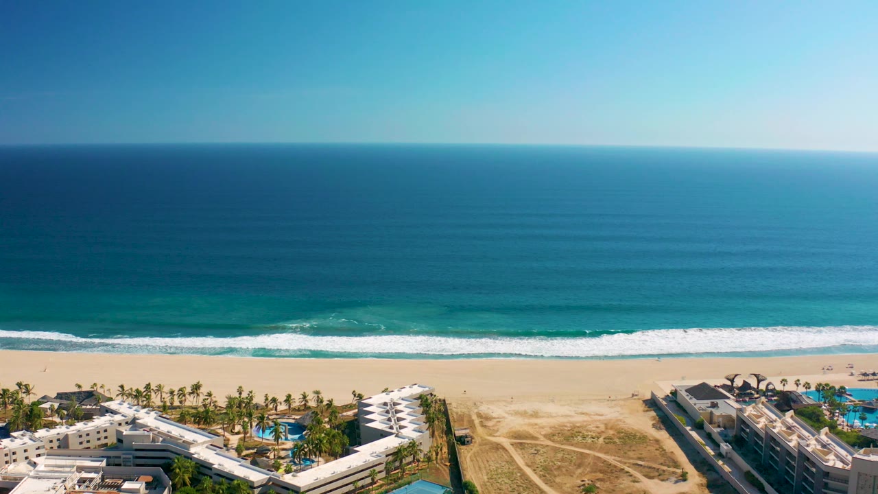Wide aerial frame over San Jose del Cabo, Mexico, showing resort buildings along the lower edge, open sand, bright surf lines, deep blue water, and no visible people.