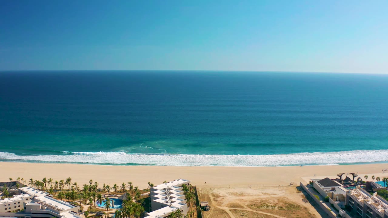 San Jose Del Cabo aerial view with the horizon high in frame, waves breaking across the beach, and white resort structures and pools along the lower edge with no visible people.