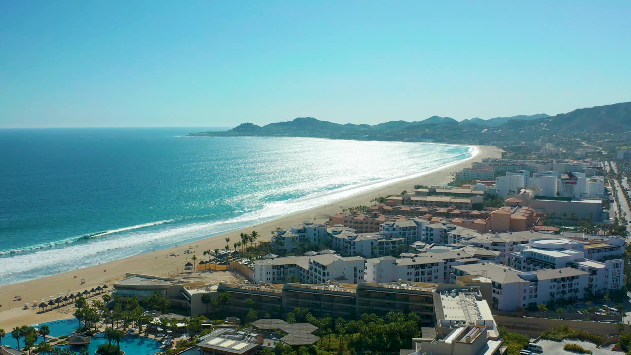 Aerial view in San Jose del Cabo, Mexico, with bright sun glare across the ocean, surf bands meeting the beach, resort buildings along the lower frame, mountains in back, and no visible people.