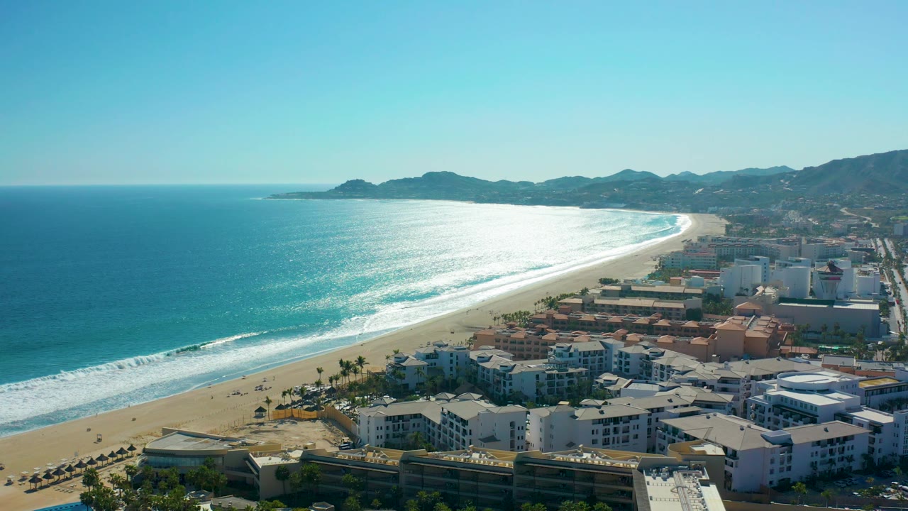 Mid-clip aerial view over San Jose del Cabo, Mexico, with foamy surf rolling toward the sandy beach, resort buildings at the bottom edge, mountains beyond the coast, and no visible people.