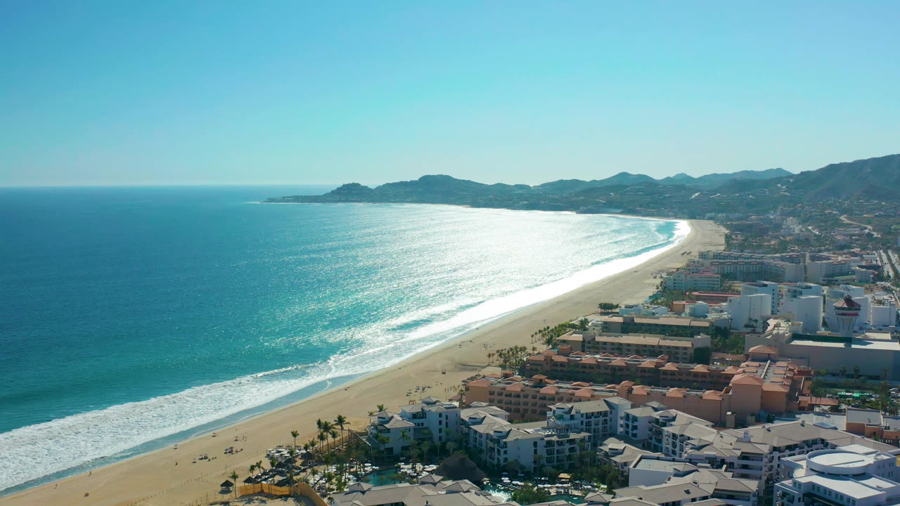 Aerial view over San Jose del Cabo beach as white surf reaches the sand, with waterfront buildings in the lower frame, mountains in the distance, clear sky, and no visible people.