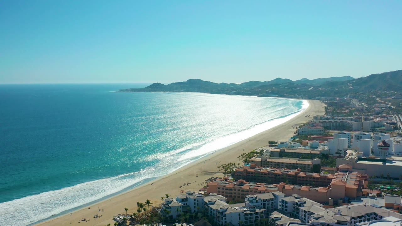 San Jose del Cabo aerial coastline view with white waves rolling toward the sand, low resort-like coastal buildings below, distant hills and mountains, clear sky, and no visible people.