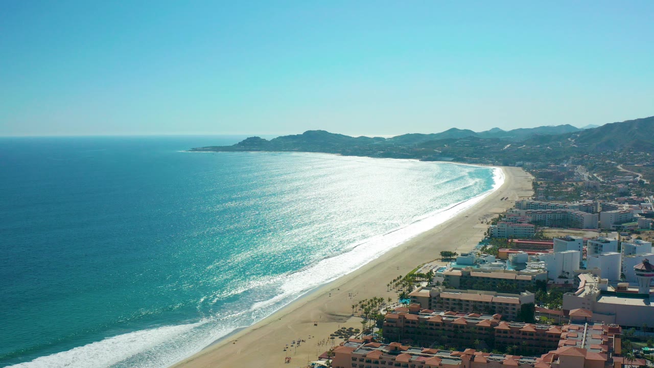 Opening aerial frame over San Jose del Cabo coastline with blue ocean at left, bright surf bands on the beach, buildings at lower right, distant mountains, and no visible people.