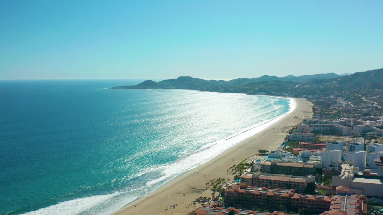 San Jose del Cabo aerial coastline with intense reflected sunlight on the ocean, pale sand below the surf, clustered buildings near the lower right edge, hills in the distance, and no visible people.