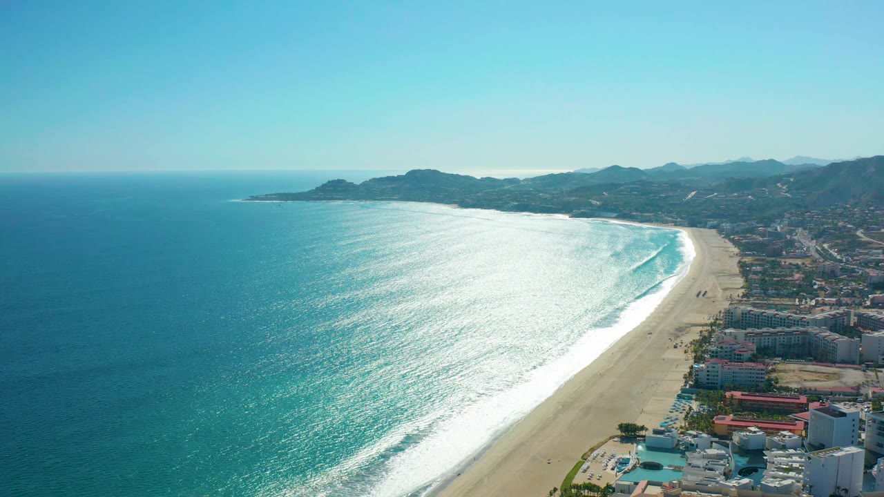 Aerial frame from San Jose del Cabo, Mexico showing sunlit ocean glare across rolling waves, a long sandy beach curving toward distant hills, coastal buildings at right, and no visible people.