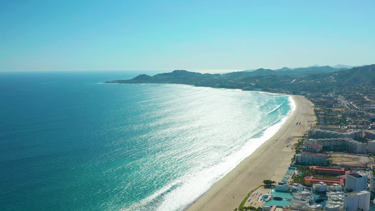 Wide aerial view in San Jose del Cabo, Mexico with open blue ocean, a broad band of white surf, sandy shoreline, coastal development on the right, mountains beyond, and no visible people.