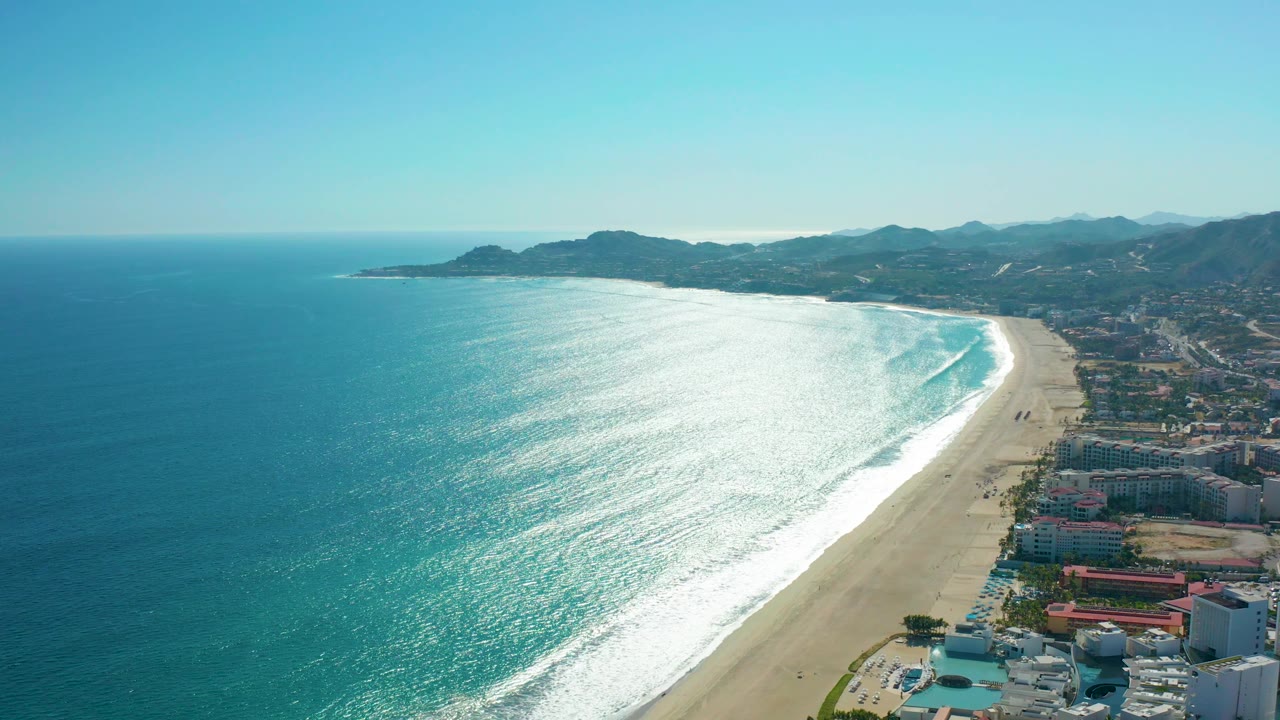 Aerial view over San Jose del Cabo, Mexico where white wave lines advance toward the long sandy beach, low coastal buildings sit near the lower right, hills rise in the background, and no visible people appear.