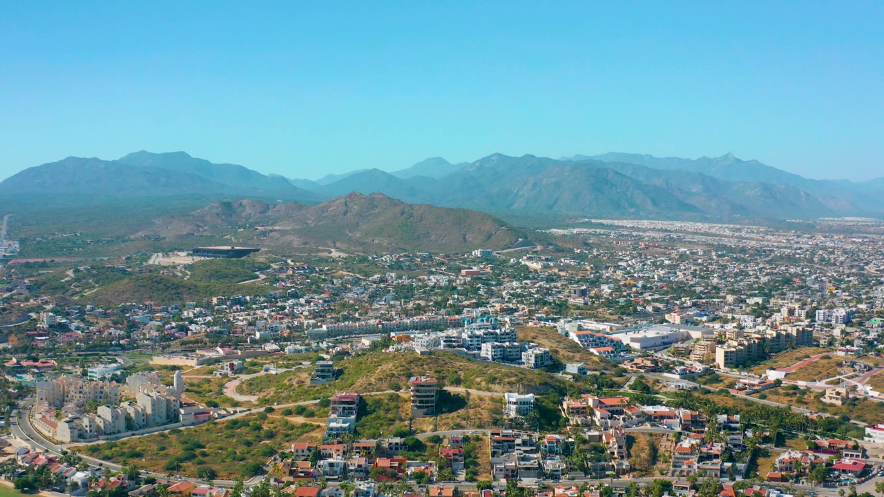 Opening aerial view over San Jose del Cabo with hillside roads and buildings in the foreground, a dense city grid beyond, distant mountains, clear blue sky, and no visible people.