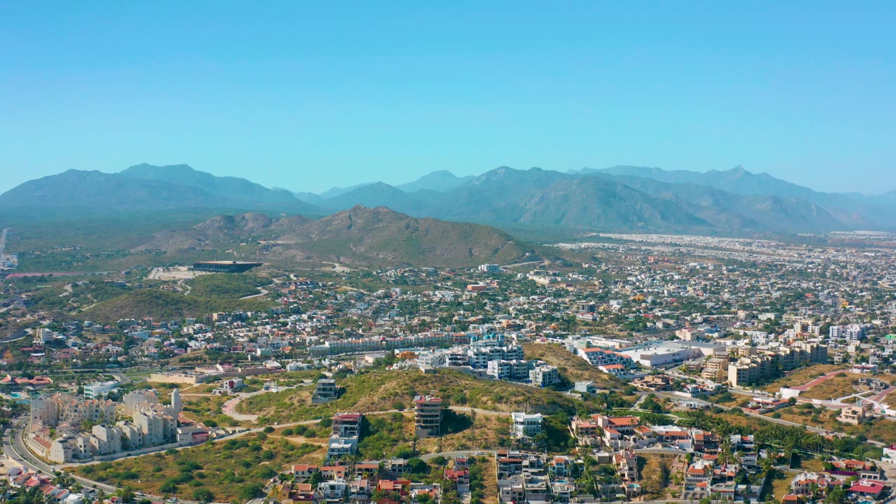 A high aerial frame shows San Jose del Cabo spread below, with a curving road and hillside homes in the foreground, larger urban blocks in the distance, mountains on the horizon, and no visible people.